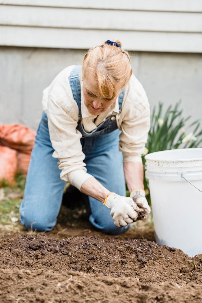 Elderly woman wearing gloves and overalls tending garden soil outdoors.