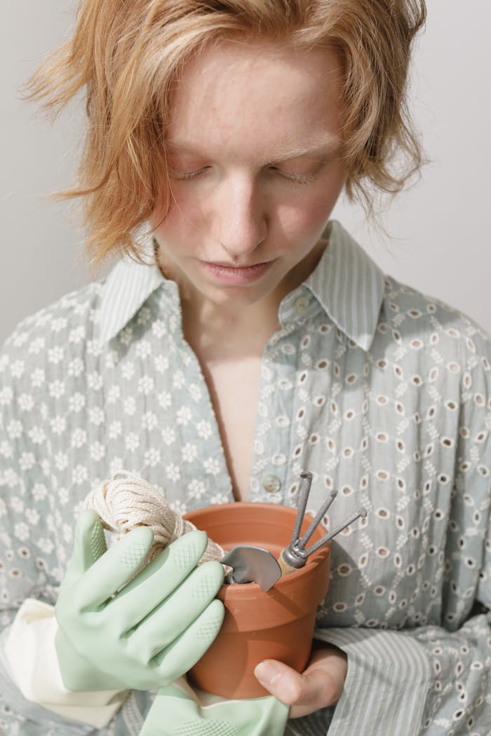 A contemplative young woman holding a clay pot with gardening tools and string indoors.